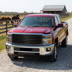 Red Chevy Silverado 2500/3500 with a satin black horizontal Stripes grille, parked on a gravel ranch lane beside a wooden horse corral.