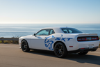 White Dodge Challenger with vivid blue Hellcat skull quarter panel graphic and bold blue SXT lettering, close three-quarter rear view on a coastal highway with Pacific Ocean in the background at golden hour