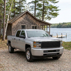 Silver Silverado HD with a satin black hex-pattern grille, parked beside a cedar cabin overlooking a lake.