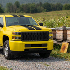 Yellow Silverado HD with black racing stripes and a satin black honeycomb grille, parked beside wooden beehive boxes.
